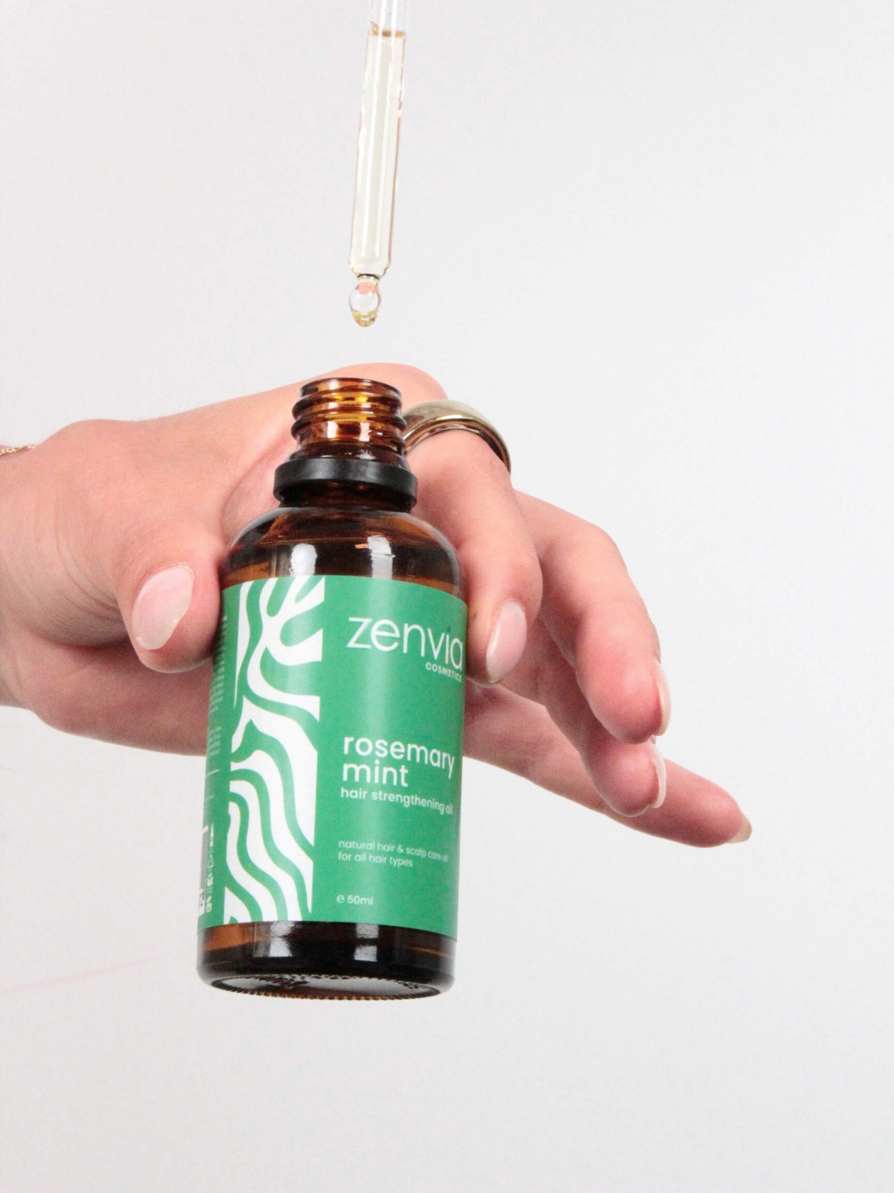 Hand holding a Zenvia rosemary mint hair strengthening oil bottle with a white background