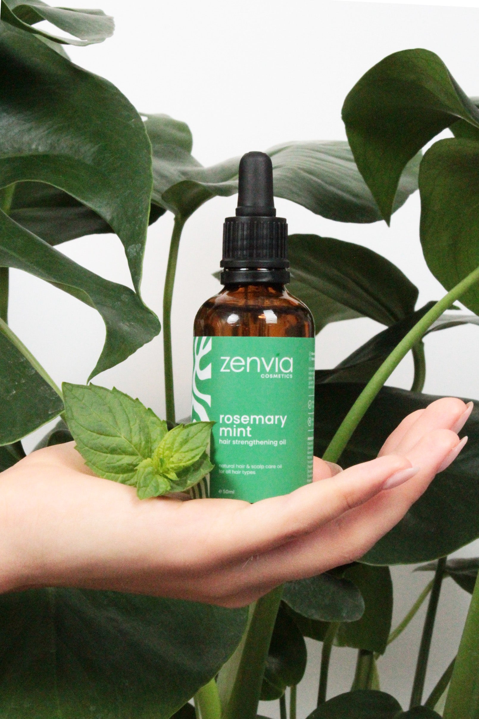 Bottle of Zenvia rosemary mint oil held in a hand with green leaves in the background