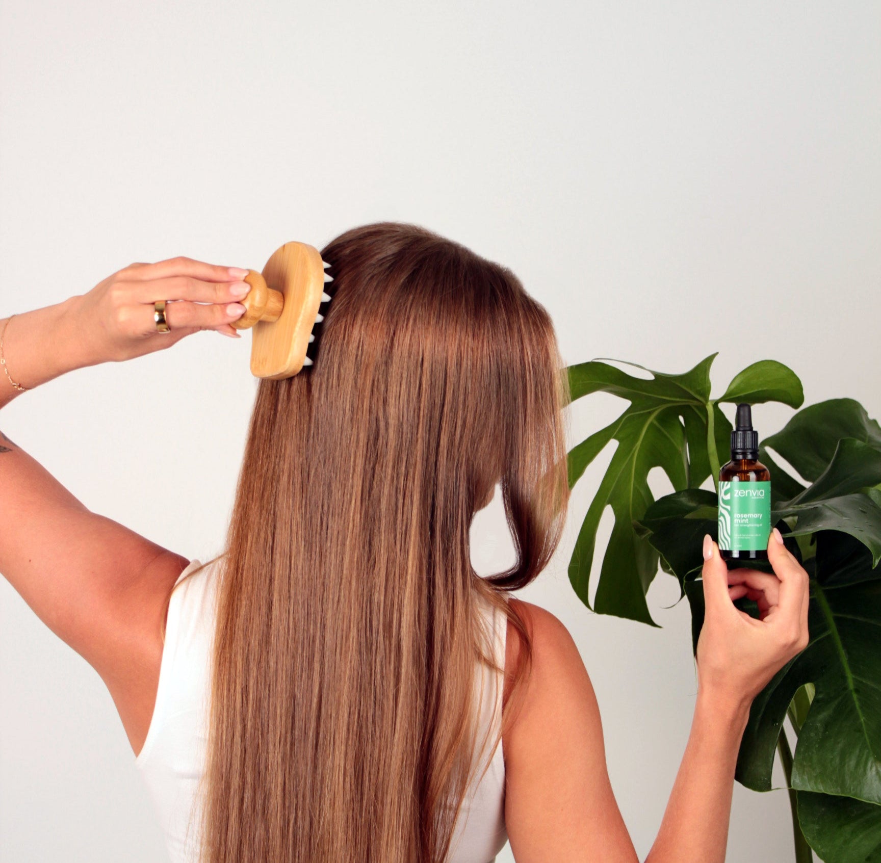 Woman with long hair using a wooden comb and holding a bottle of Zenvia hair care product against a white background.